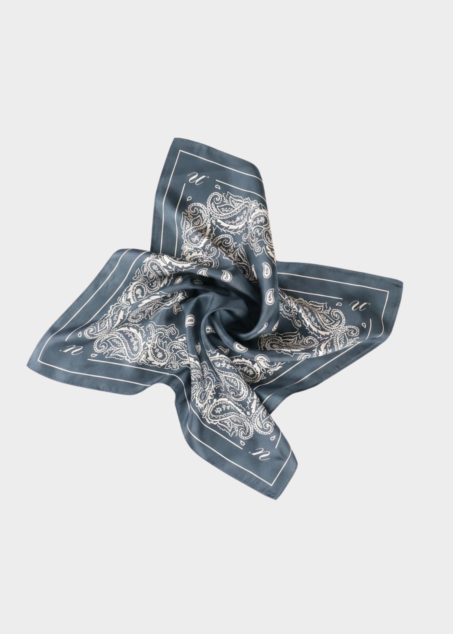 Noted Antwerp Alice scarf in denim blue. Rectangular scarf with with paisley print on denim blue 100% silk scarf.
A square bandana scarf with a muted blue-gray base and an intricate white paisley pattern, styled in a central swirl on a light gray background.