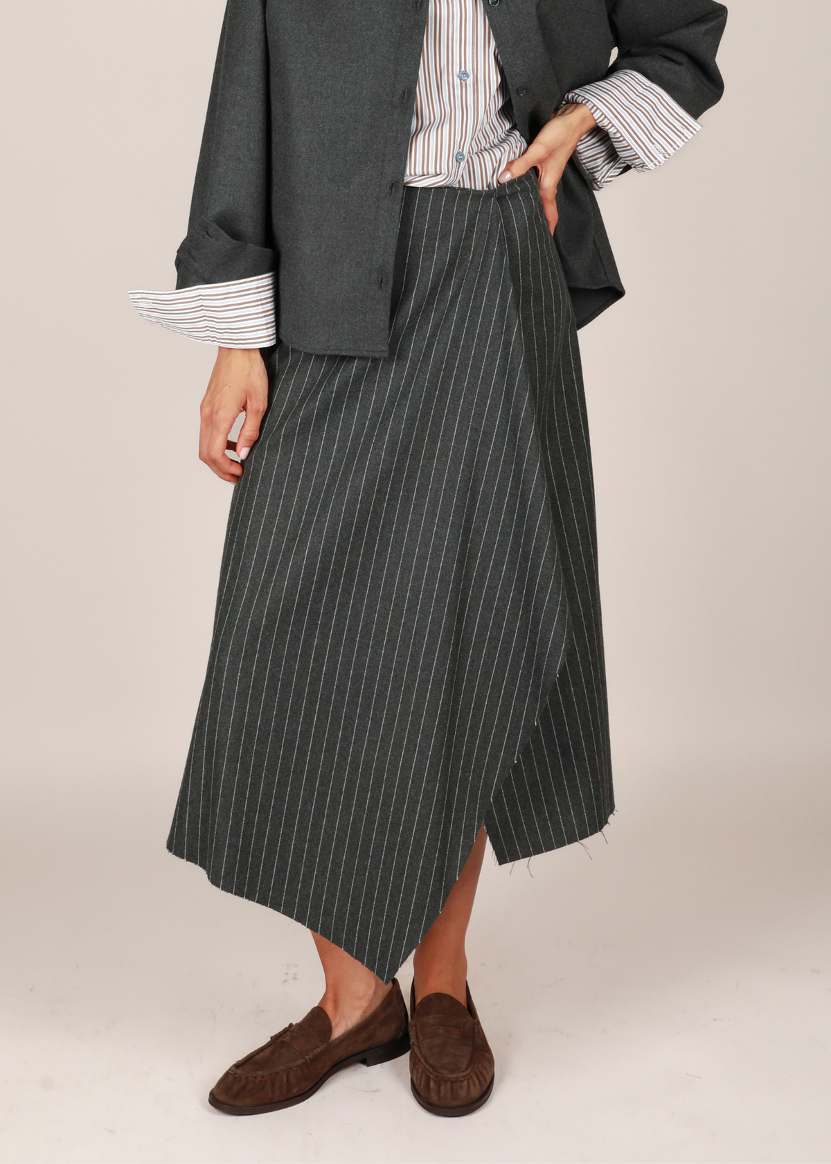 Noted Antwerp Dodo wrap skirt. A model wearing a dark gray pinstriped wrap skirt, styled with a striped shirt, a dark gray blazer, and brown suede loafers, against a plain beige studio background.