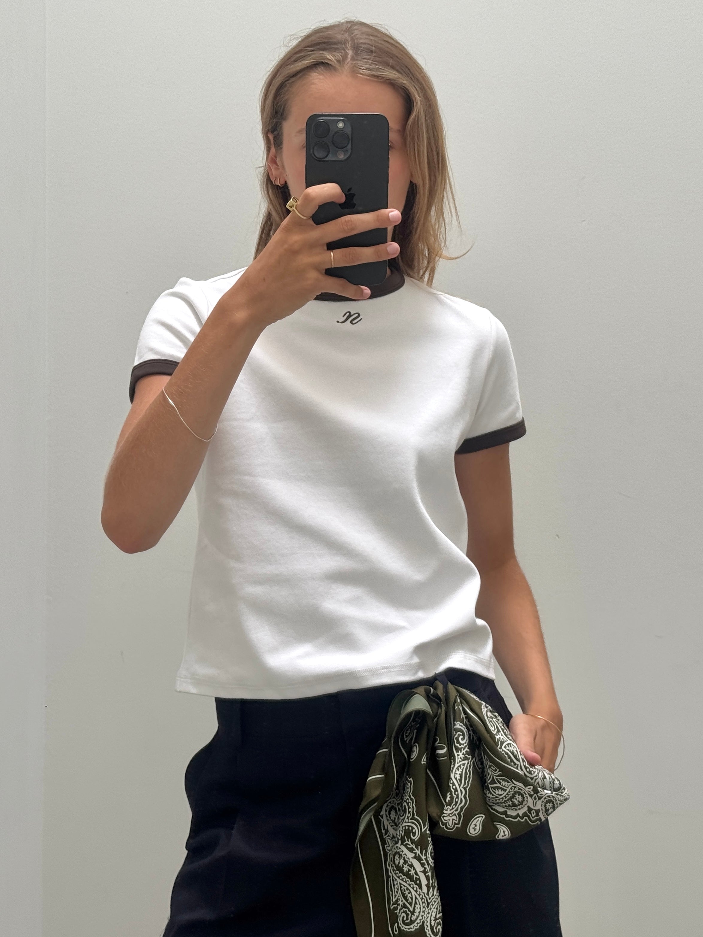 A woman takes a mirror selfie wearing a white short-sleeve t-shirt with khaki ringer trim on the collar and sleeve cuffs paired with the khki Noted antwerp alice scarf.
