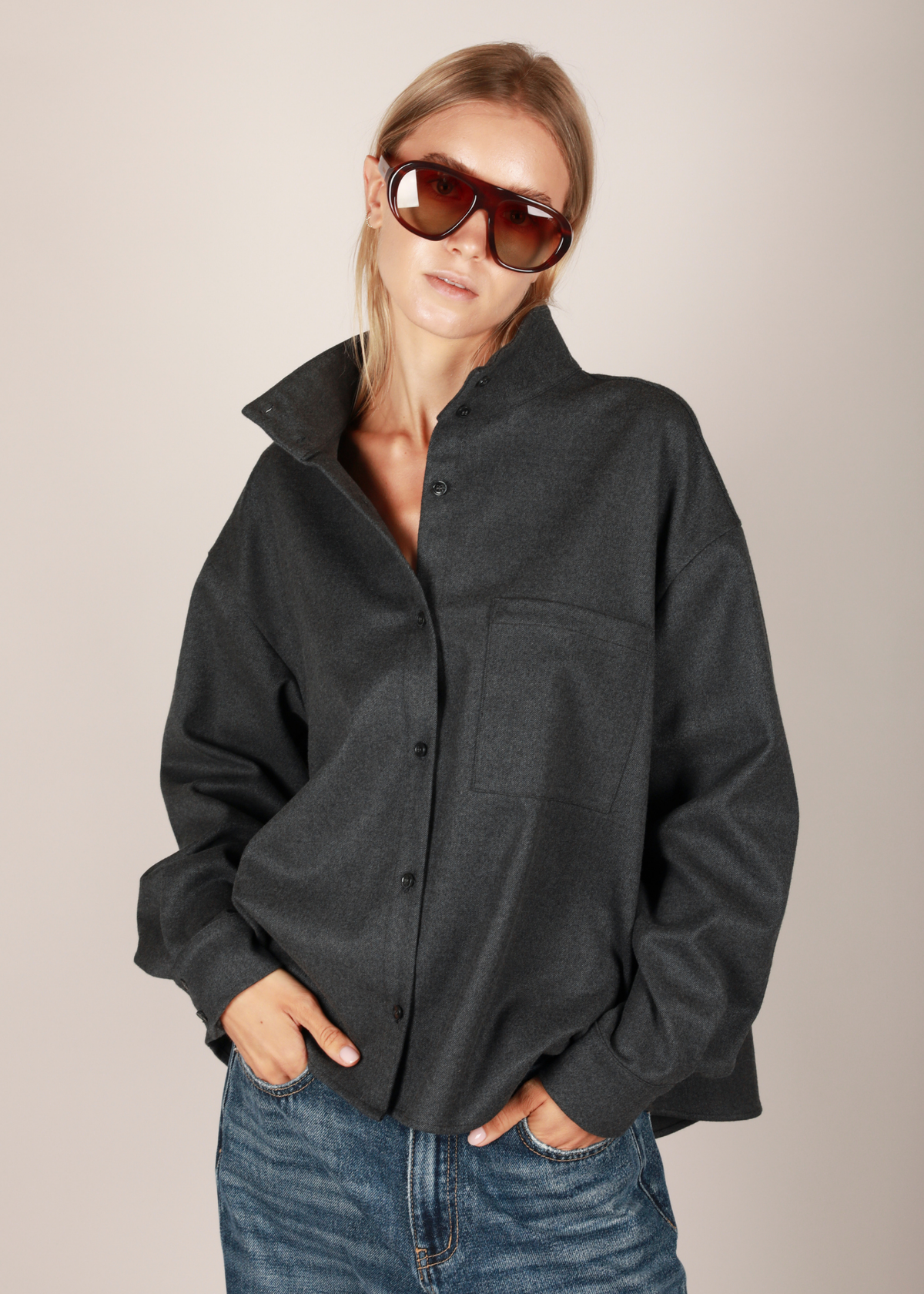 NOTED didi shirt oversized dark charcoal gray button-up shirt with a high collar and chest pocket, paired with blue jeans and sunglasses, against a solid light beige background.
