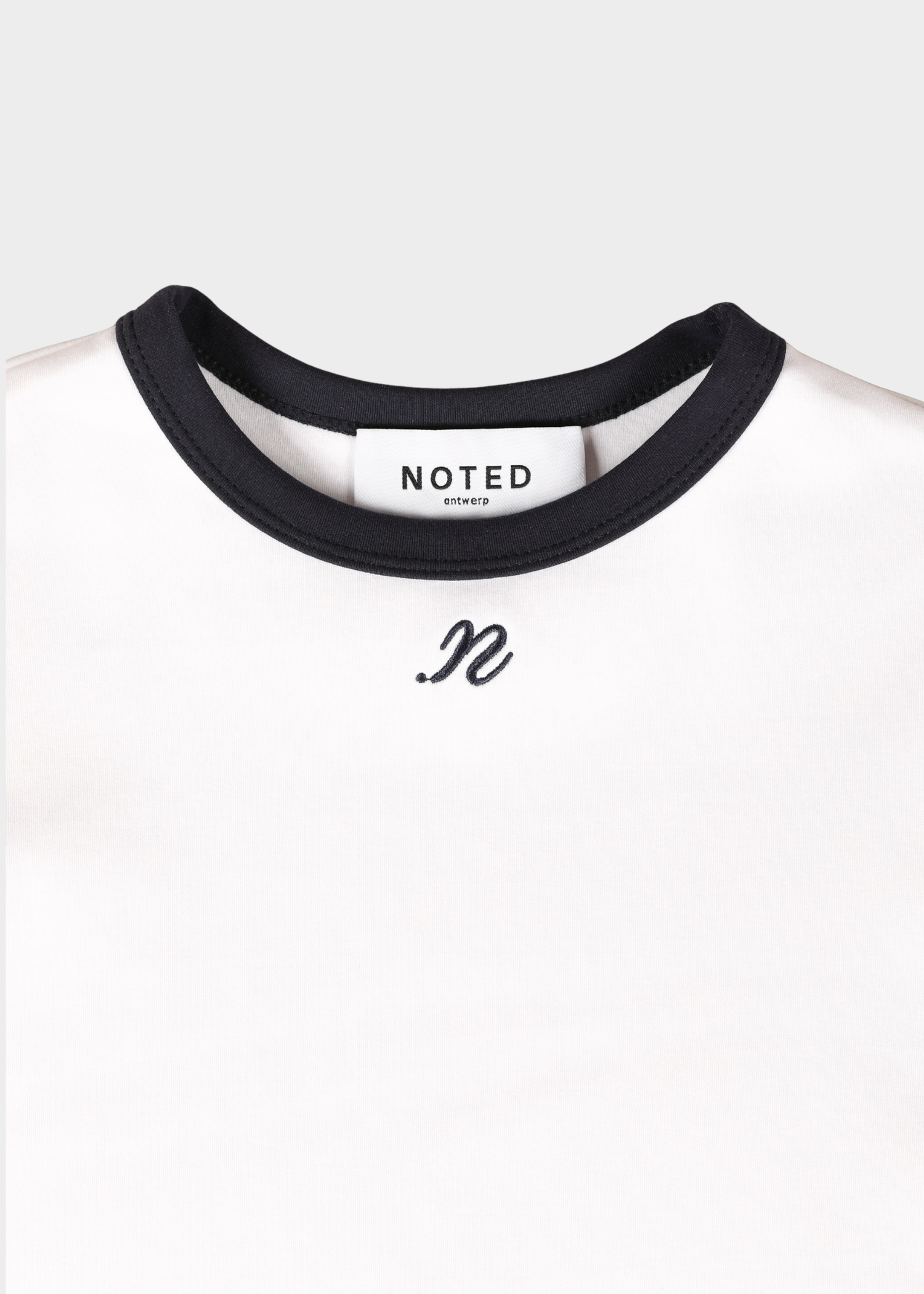 Close-up of a white ringer t-shirt with a black ribbed crew neck and a black embroidered script logo on the chest. The neck label reads 'NOTED'.