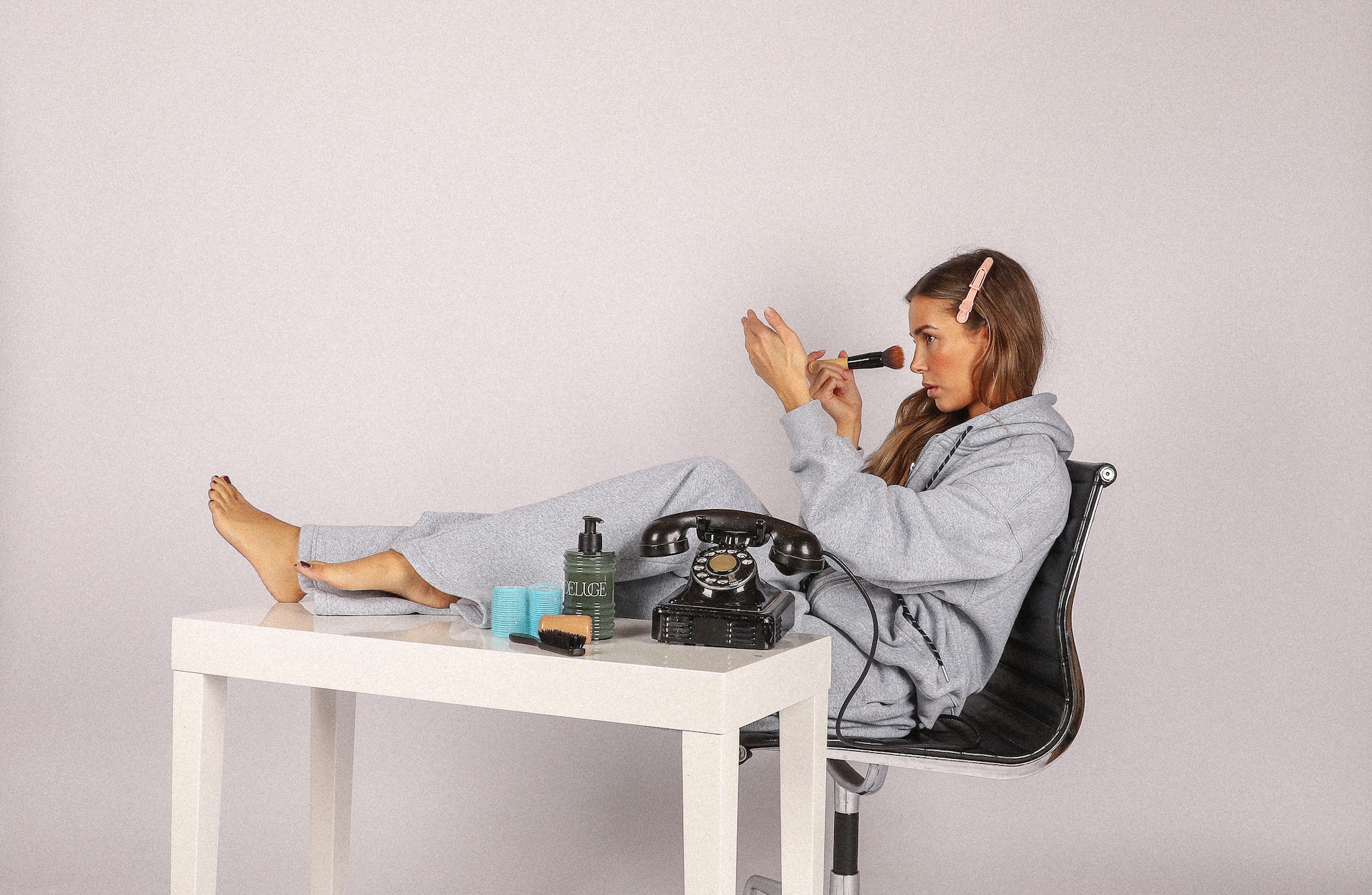 Woman sitting on a chair with a phone and makeup items, wearing a gray hoodie.