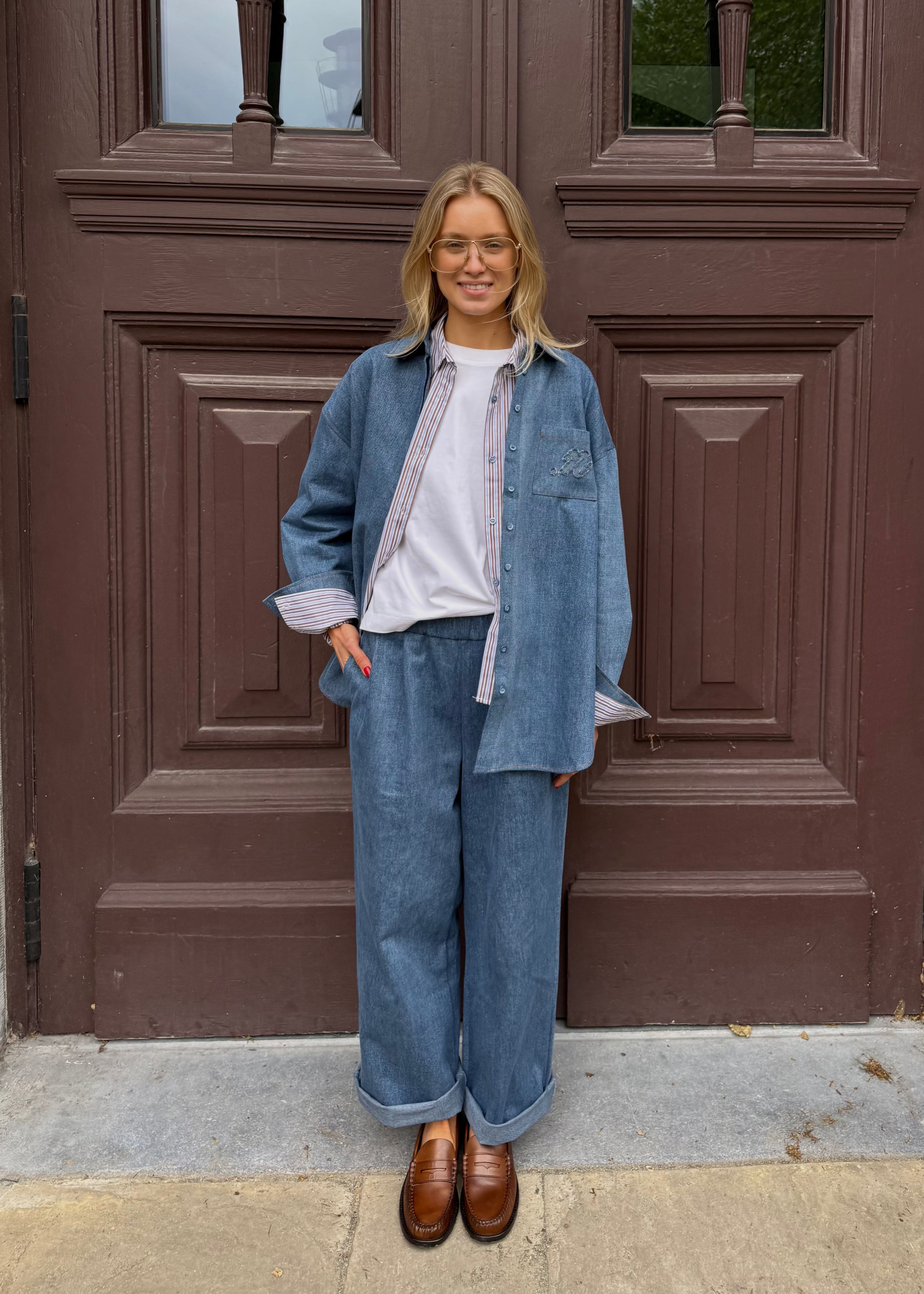 A woman stands in front of a large brown wooden door wearing a matching blue denim outfit consisting of a button-up shirt and wide-leg pants, layered over a white t-shirt and finished with brown leather loafers.