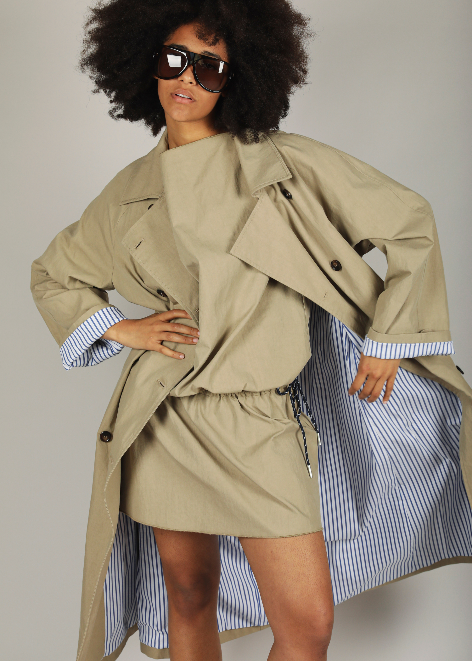 Person wearing a beige trench coat over a striped dress with sunglasses on a plain background