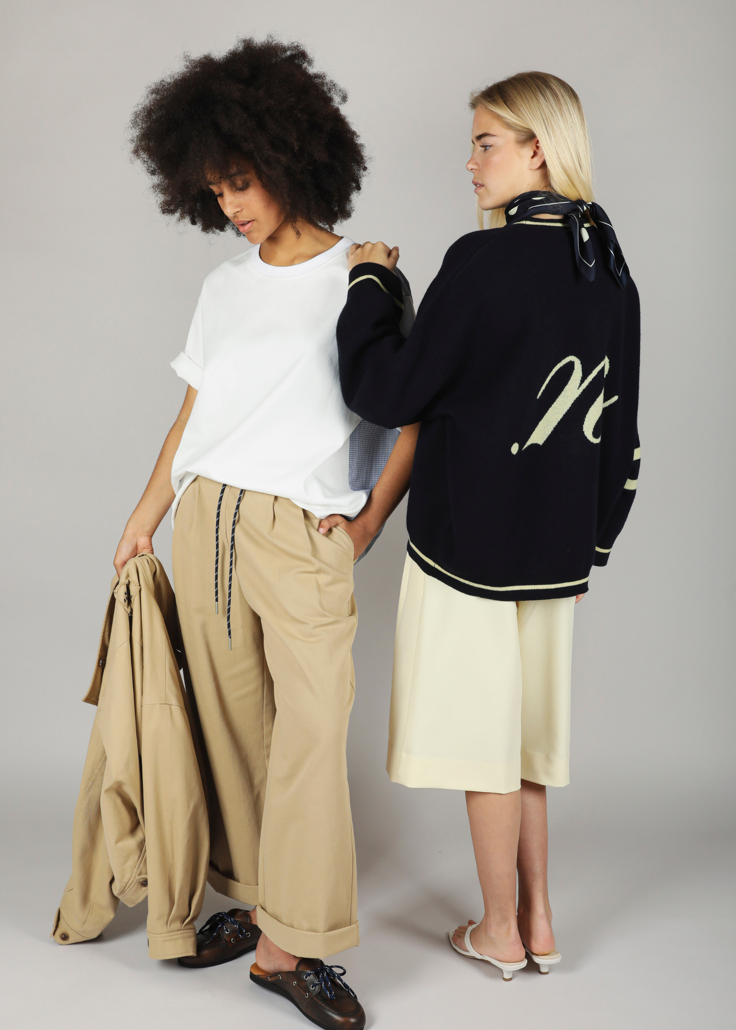 Two women standing close together, one wearing a blue benny cardigan from noted antwerp and beige pants, the other in a white shirt and beige pants. the whole outfit is noted antwerp 