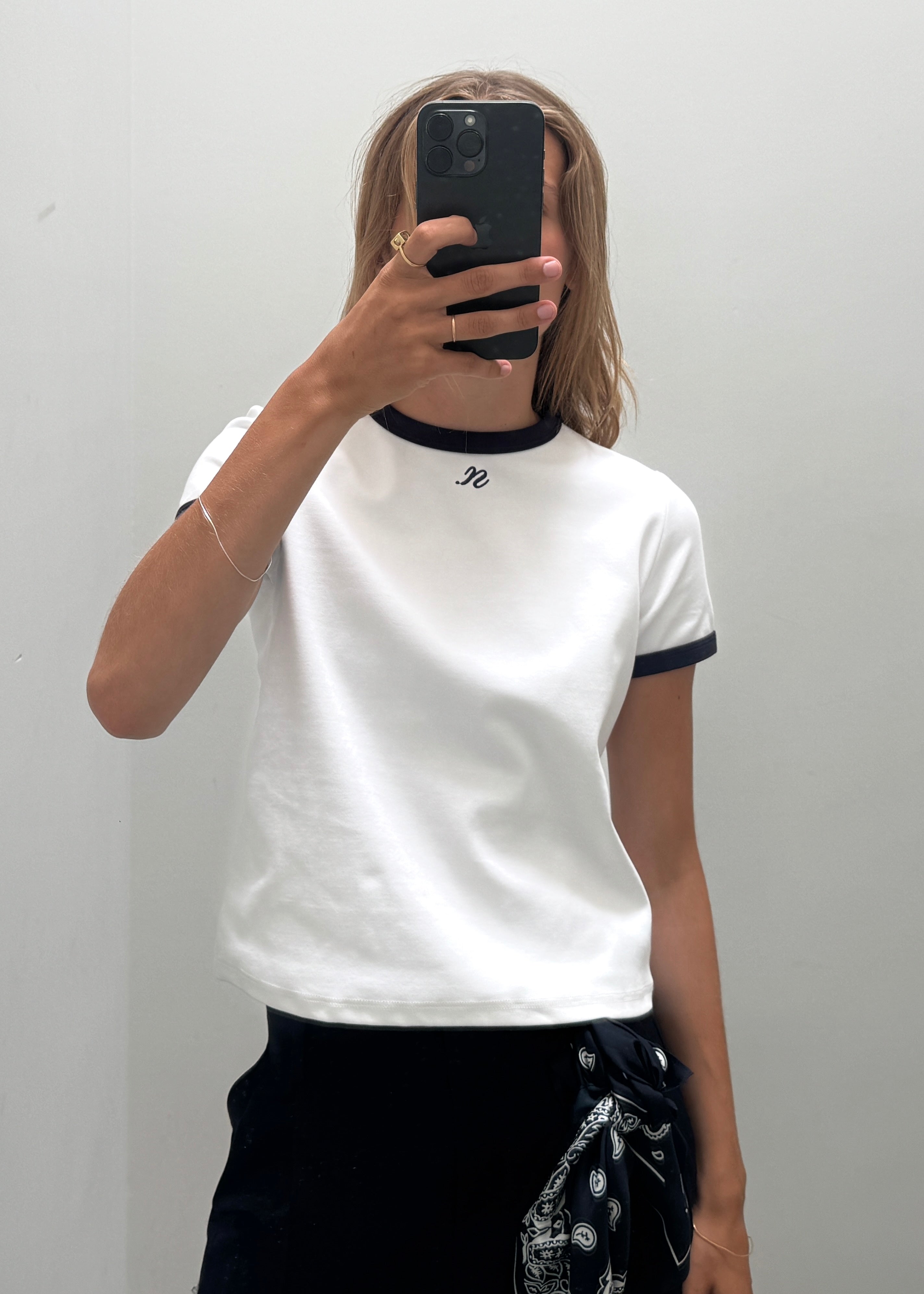 A woman taking a mirror selfie wearing a white ringer t-shirt with dark blue trim at the collar and sleeve cuffs and embroidered noted antwerp logo on the front chest.