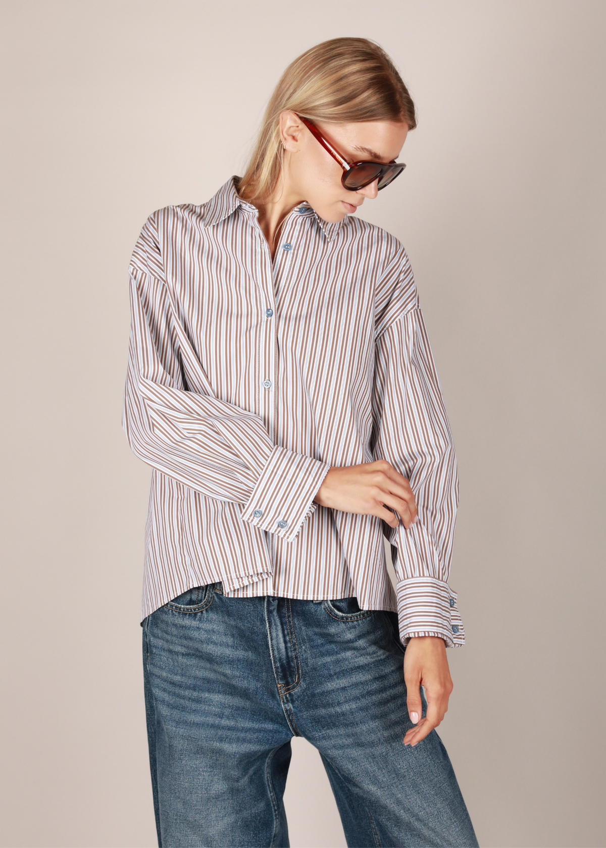 NOTED ANTWERP Anni shirt in blue brown striped pattern. A woman wearing this longsleeve button-up shirt with thin vertical red and white stripes, paired with blue jeans and red-framed sunglasses, posed against a solid light beige background.