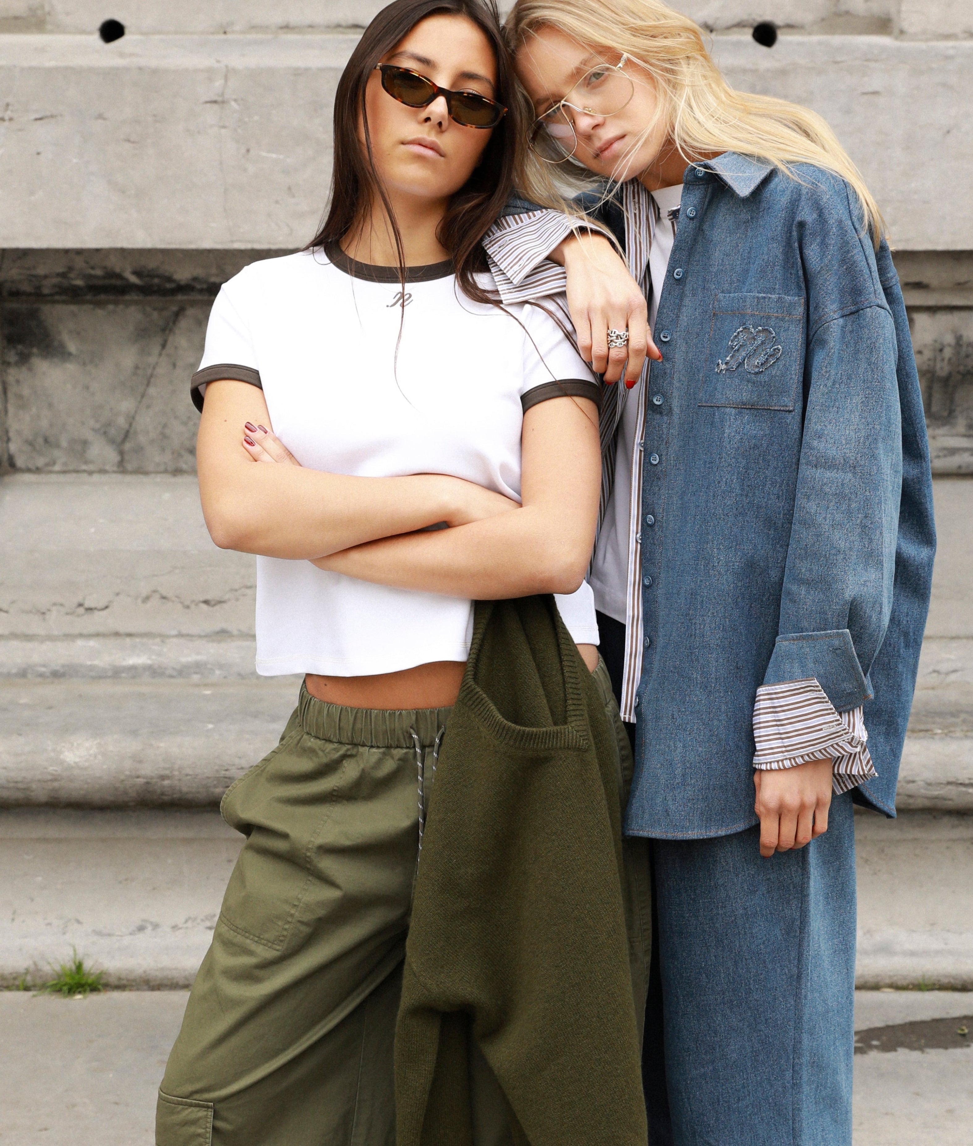 Two women posing together outdoors in front of concrete steps, showcasing a streetwear collection. One wears a white ringer t-shirt, olive green cargo pants, and tan sneakers, while the other wears a blue denim jacket and matching wide-leg jeans with brown leather loafers.