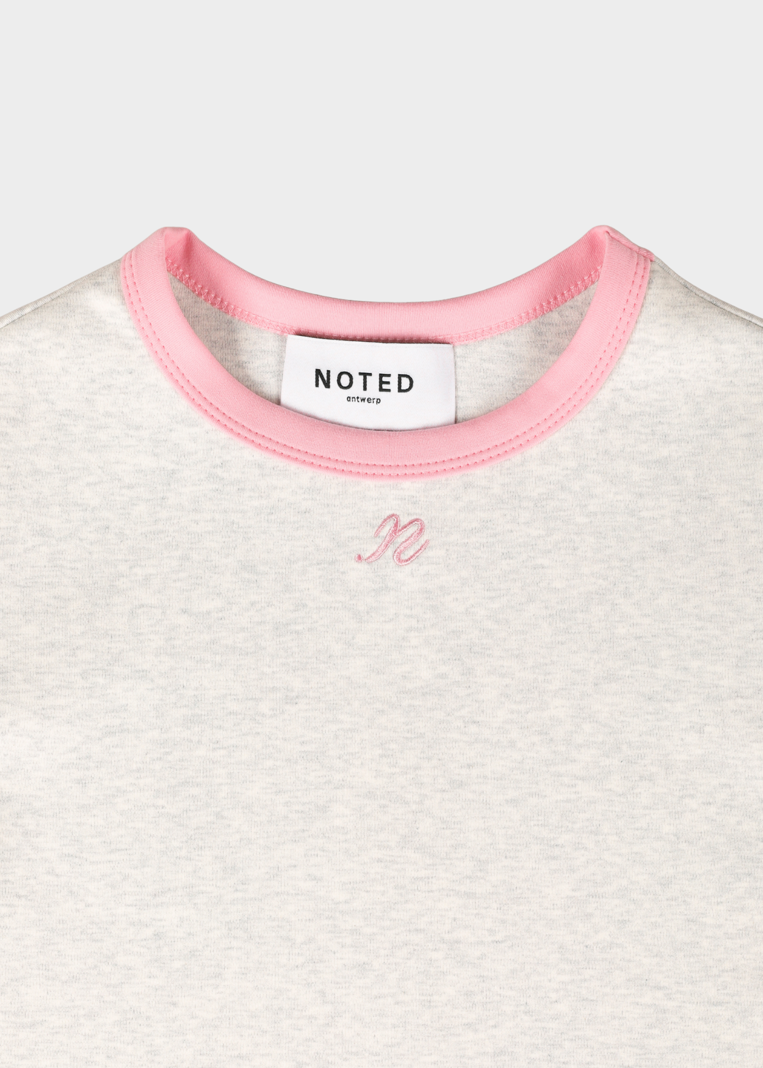 Close-up of a light heather grey t-shirt with a pink ribbed crew neck and a small pink embroidered monogram on the chest. The neck label reads 'NOTED ANTWERP'.