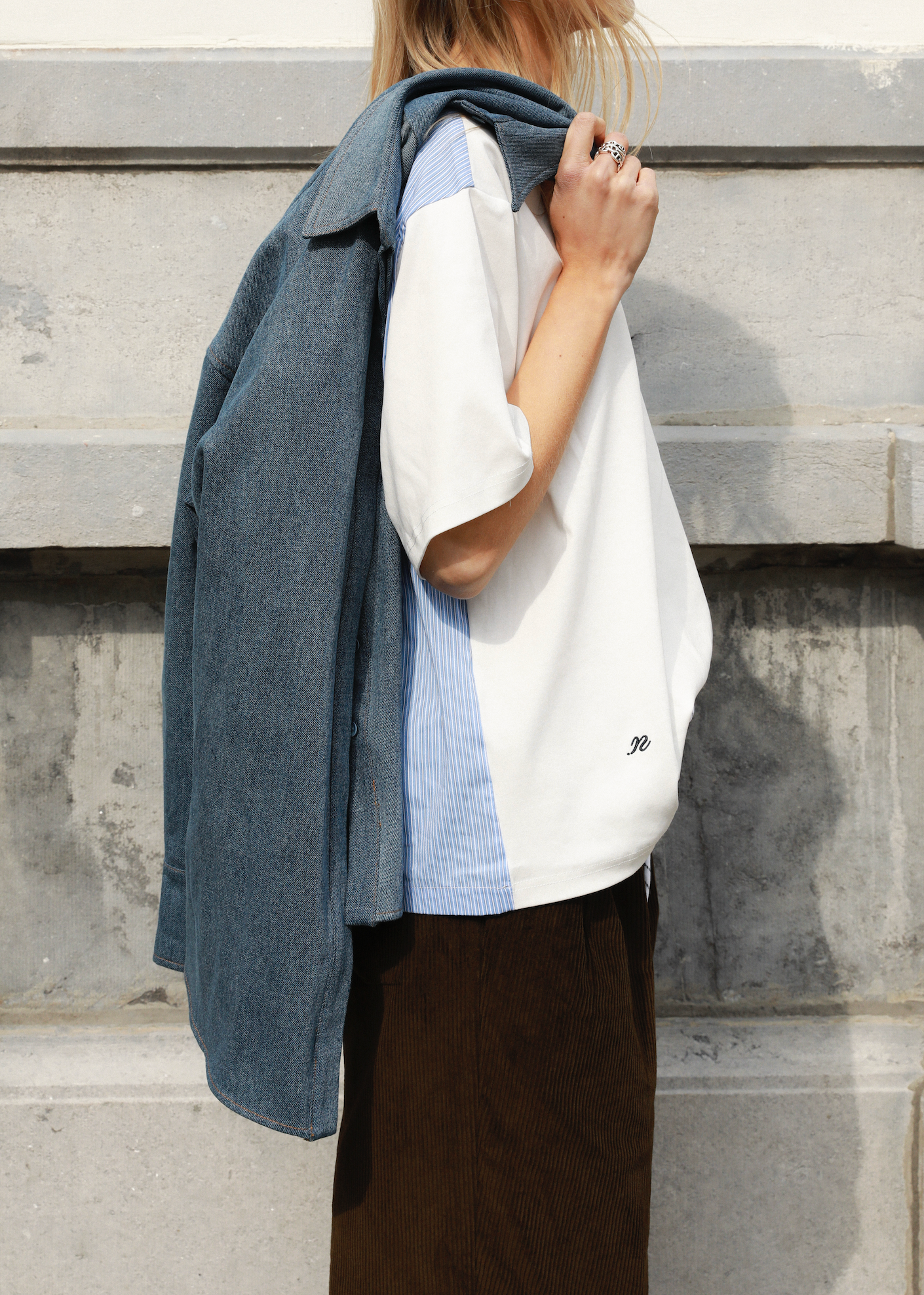 A model wearing a white short-sleeve t-shirt with light blue and navy vertical color-blocked stripes, a blue denim overshirt draped over her shoulders, and dark brown trousers, standing against a gray concrete wall.