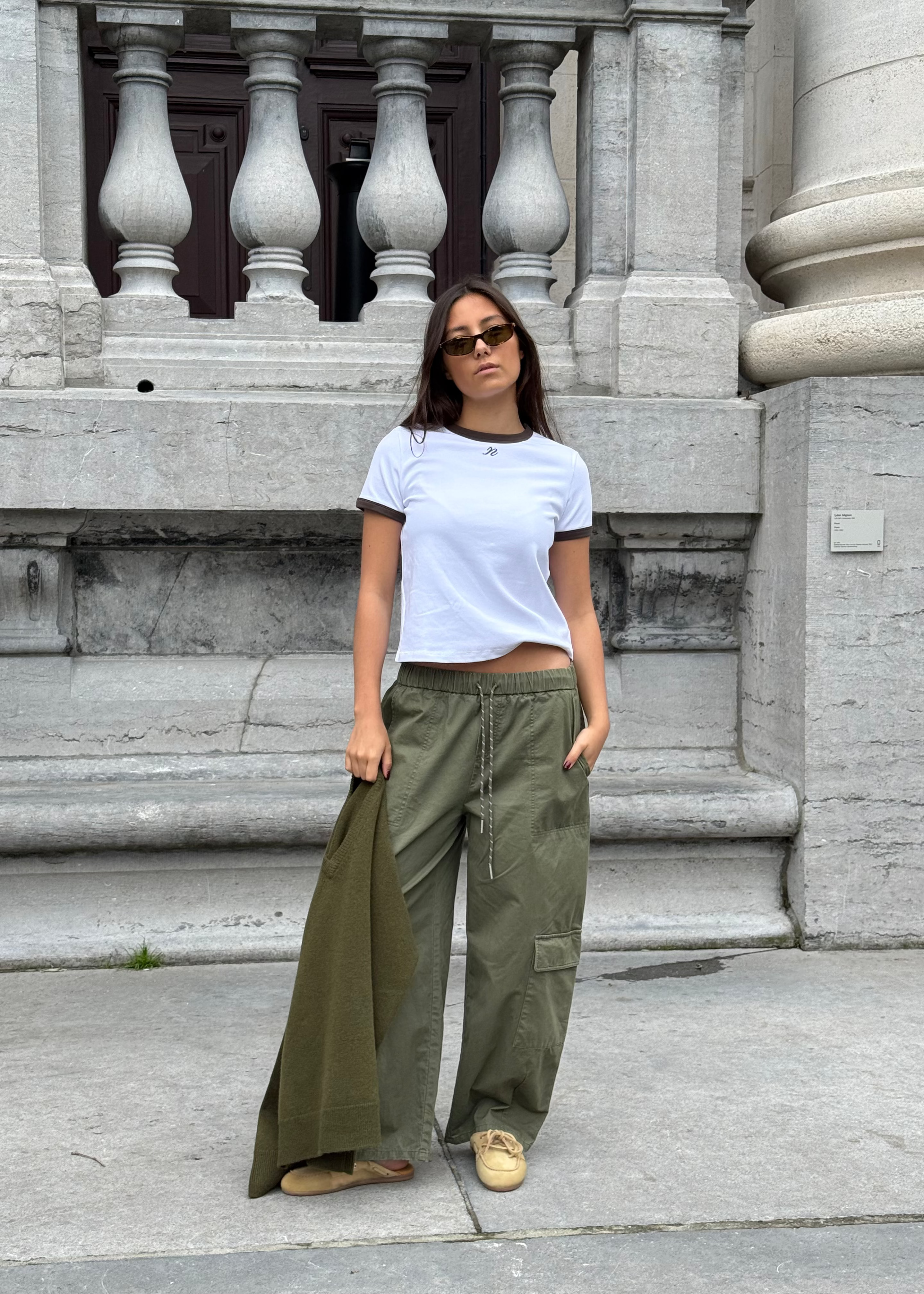 A woman stands on a stone sidewalk in front of a grand classical building, wearing a white ringer t-shirt, olive green wide-leg cargo pants, and tan sneakers.